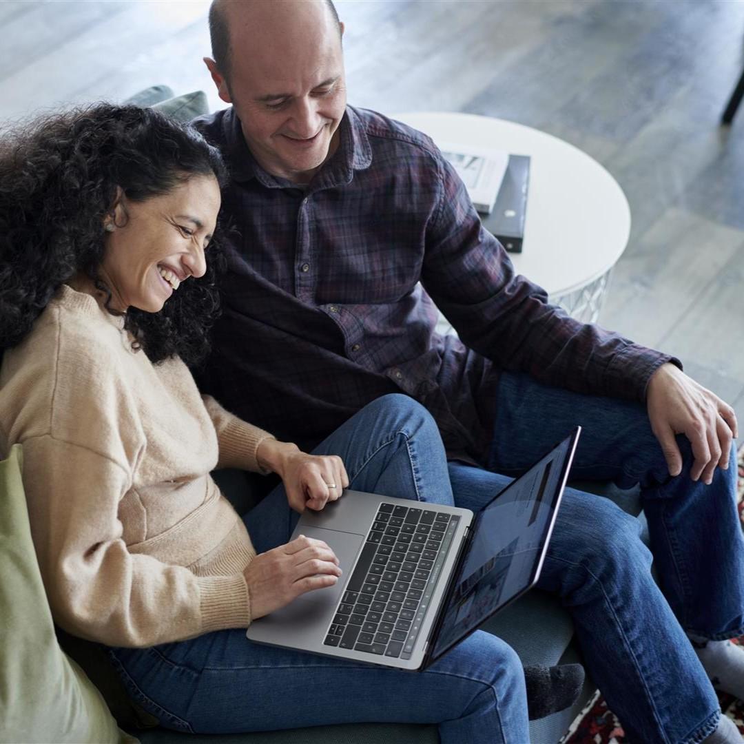A woman and a man are sitting on the sofa looking at a laptop that the woman has on her lap.