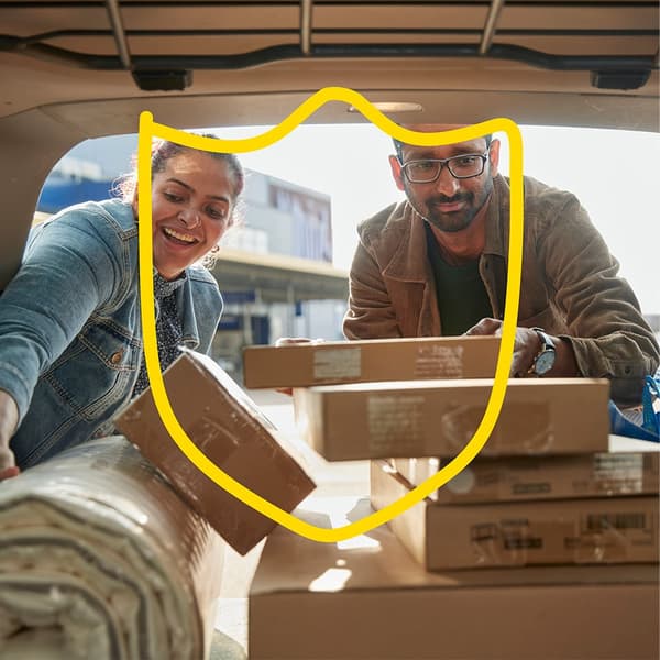 A woman and a man safely move a rug and IKEA products packed in cardboard boxes into a car. Above them is an IKEA illustration of a safety (protection) symbol.