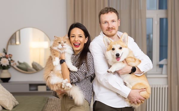 A woman and a man standing in a bright bedroom, holding a cat and a dog and posing for the camera in a cosy, warm atmosphere.