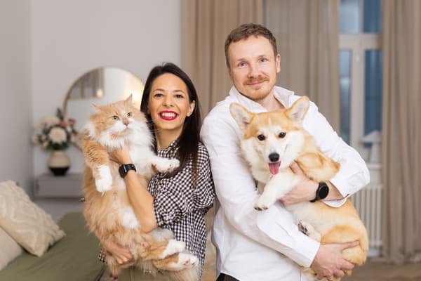 A woman and a man standing in a bright bedroom, holding a cat and a dog and posing for the camera in a cosy, warm atmosphere.