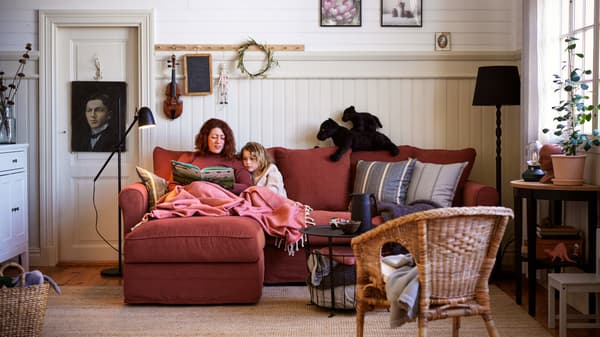 A woman and girl cosy up with a book on a light red GRÖNLID three-seater sofa with a chaise longue in a living room.
