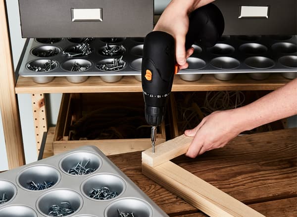 A woman at a workbench drills together a large frame, with a muffin tin filled with hardware by her side.
