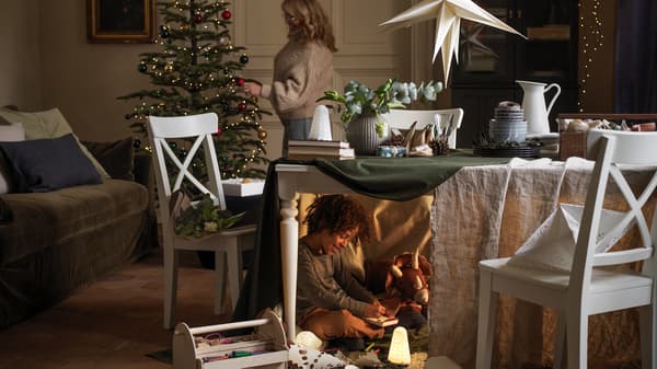 A woman decorates a Christmas tree, while under the table in front of it a boy plays, half-hidden, with his toys.
