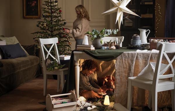 A woman decorates a Christmas tree, while under the table in front of it a boy plays, half-hidden, with his toys.