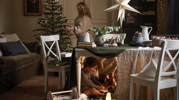 A woman decorates a holiday tree, while under the table in front of it a boy plays, half-hidden, with his toys.