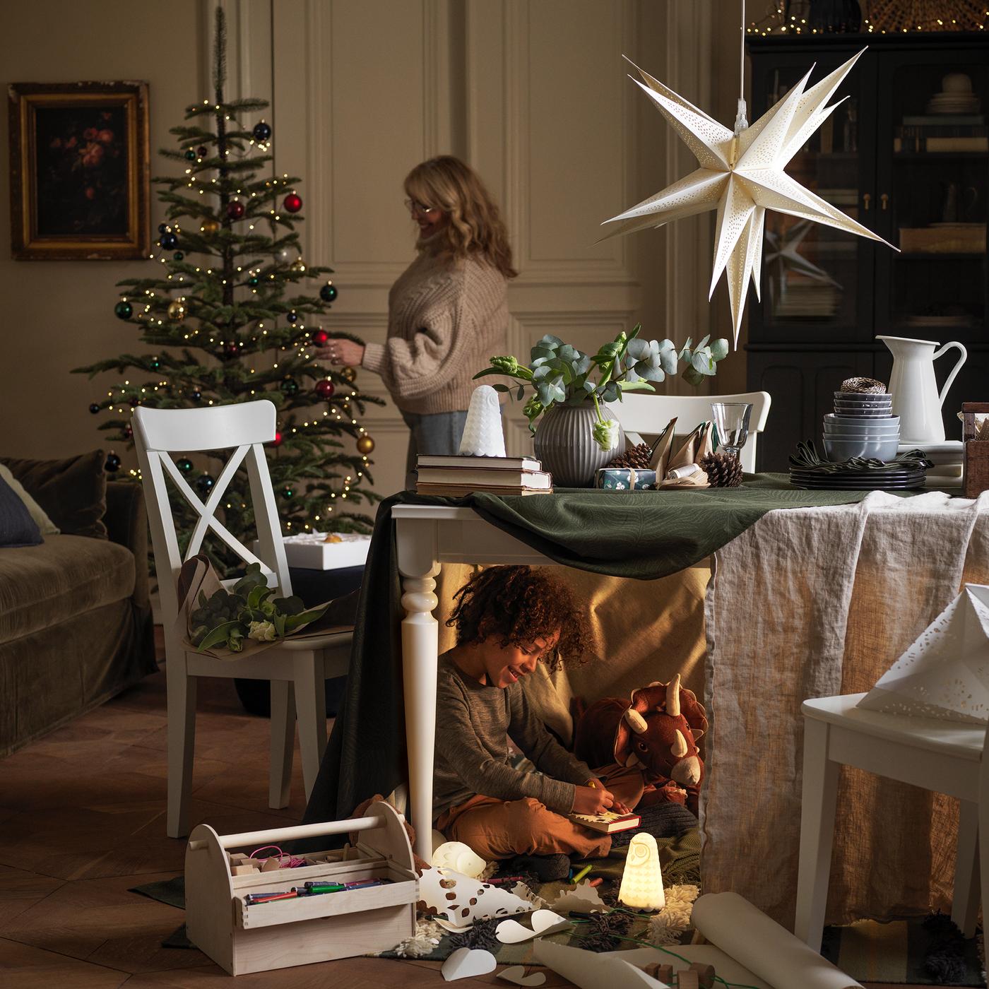 A woman decorates a holiday tree, while under the table in front of it a boy plays, half-hidden, with his toys.