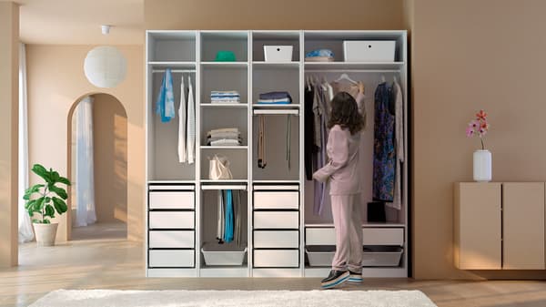 A woman hangs a dress on the clothes rail of a white, open PAX wardrobe combination in a room with light brown walls.