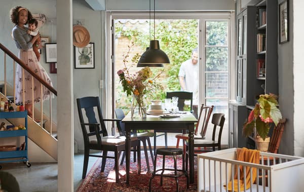 A woman holding a baby coming down the stairs into a dining area with table and chairs, with doors  out to a garden.