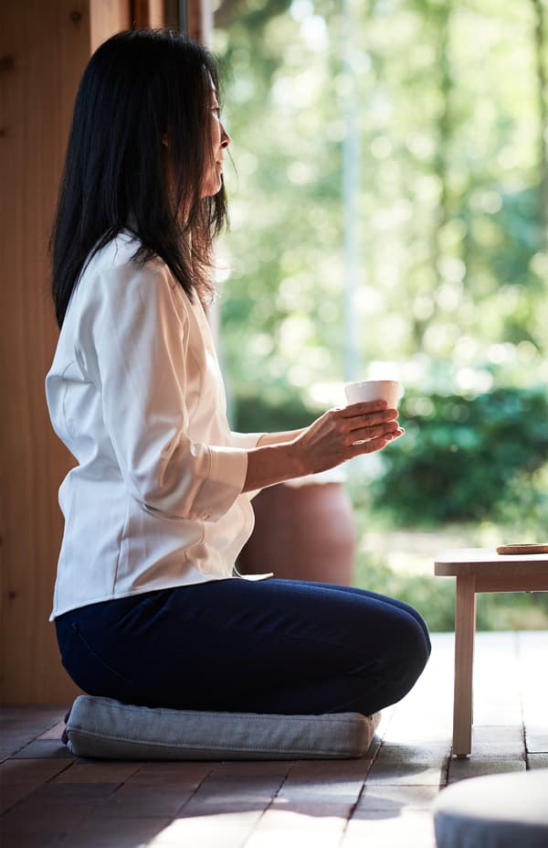A woman holding a mug in both hands sits on an OMTÄNKSAM chair cushion placed on the floor.