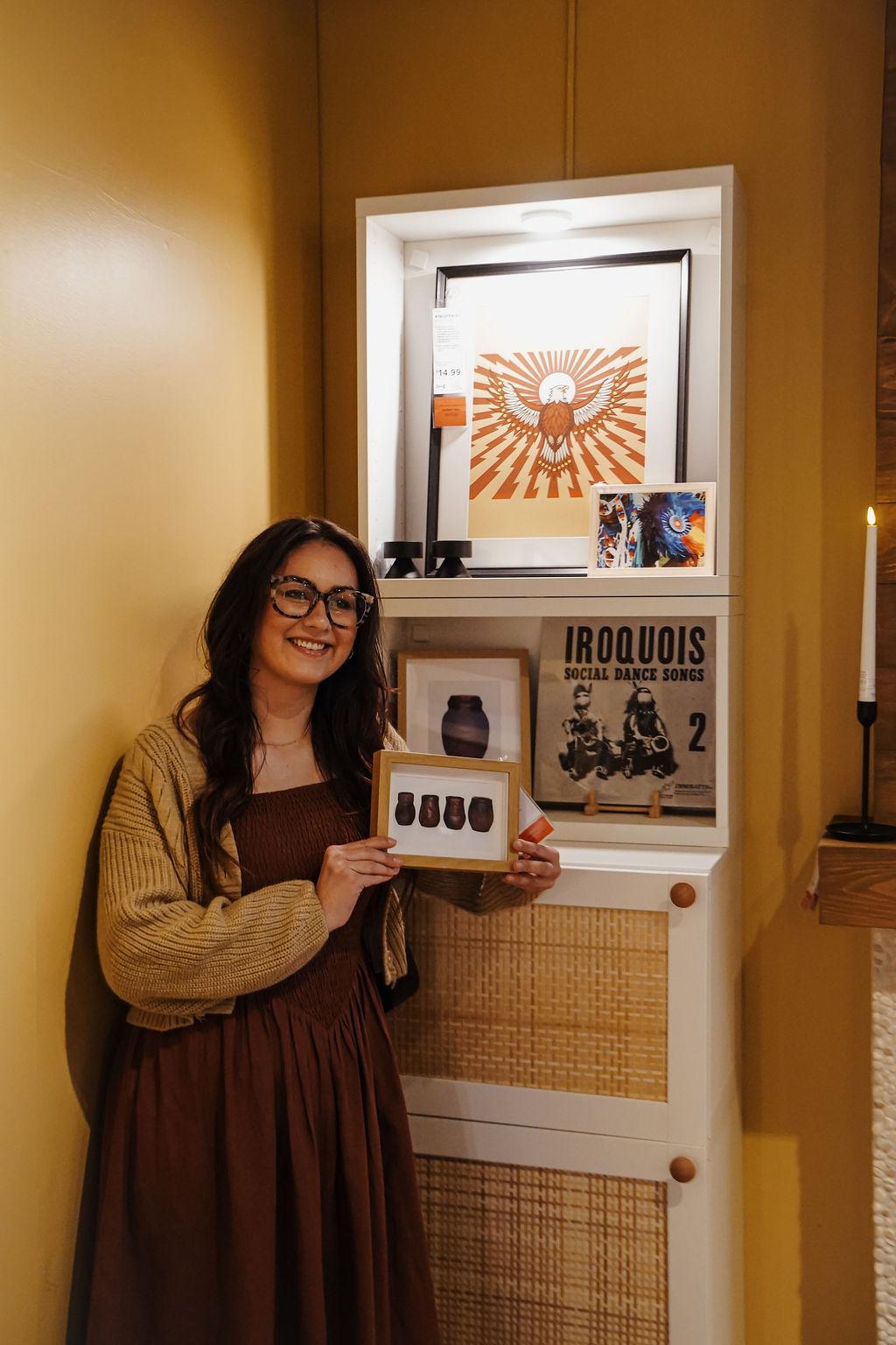 A woman holds a framed piece of art and is standing in front of other art pieces.
