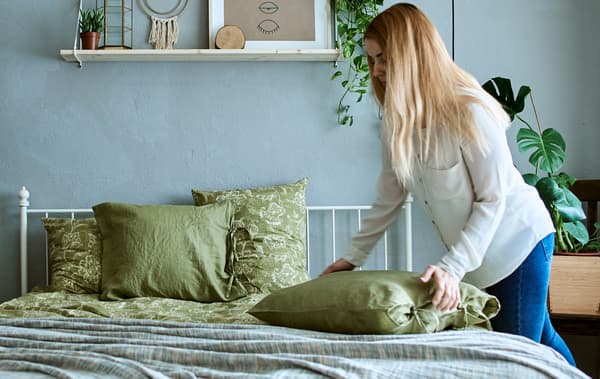 A woman in a bedroom with grey and green decor, filled with plants, plumps a pillow on a bed made up with green bedlinen.