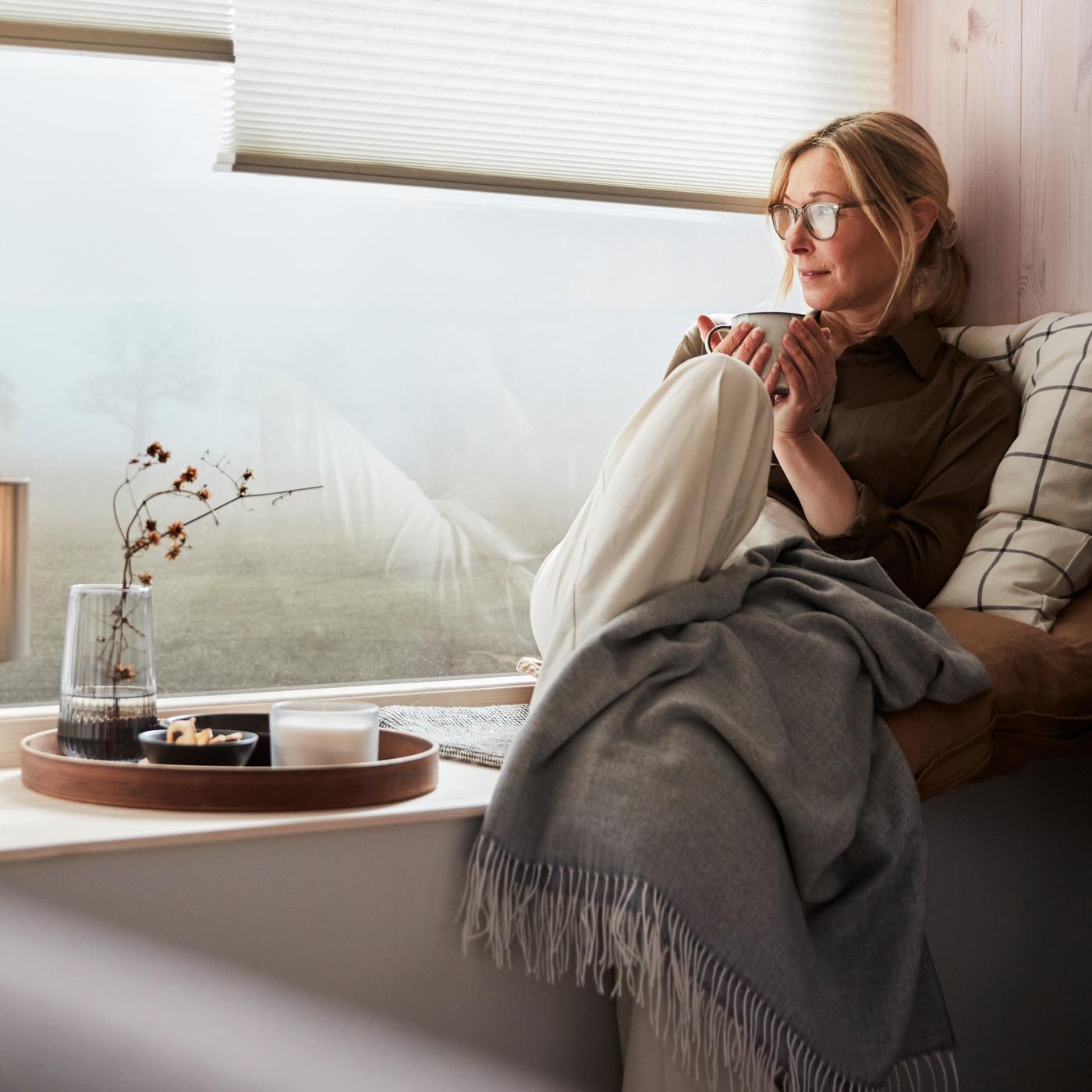 A woman in a window seat enjoying a break, with a HOLMVI throw and leaning into a cushion with a SPIKKLUBBA cushion cover.
