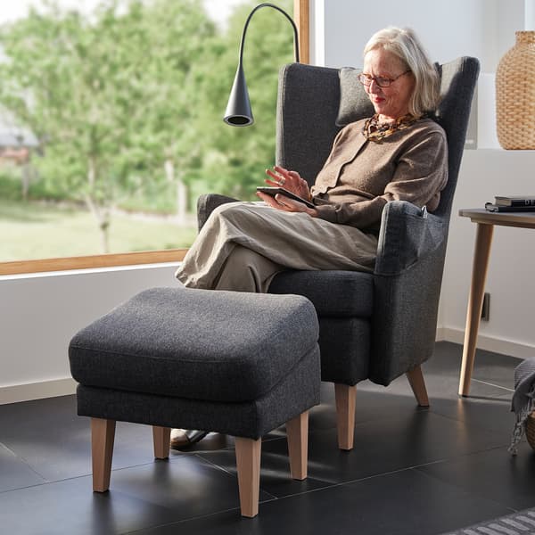 A woman in her 60s sits in an OMTÄNKSAM armchair. The armchair and footstool are placed in front of a large window.