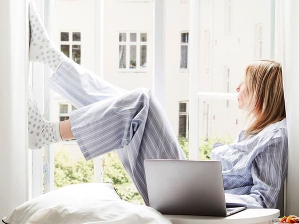 A woman in pyjamas is sitting in the window frame with a laptop beside her