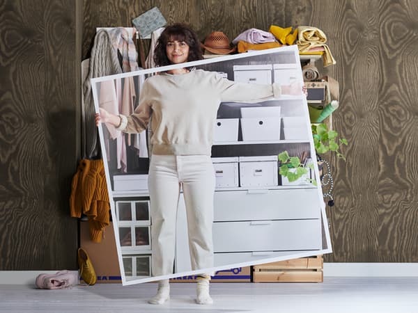 A woman in white clothing stands in front of messy shelves holding a large picture of the shelves when they've been tidied up.