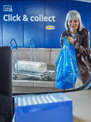 A woman is holding a blue ikea bag containing some products. Behind her, a wrapped mattress is placed on a cart. On the blue wall behind her, "click & collect" is written with white letters 