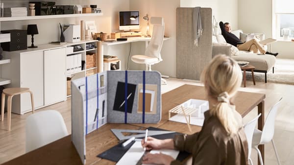 A woman is sitting at a dining table using ÖVNING desk divider to study and is looking over at a man sitting on a sofa.