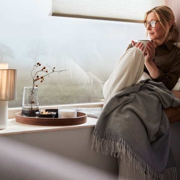 A woman is sitting in a window seat enjoying a break with a cup of tea. On the window ledge is a SYMFONISK speaker lamp.