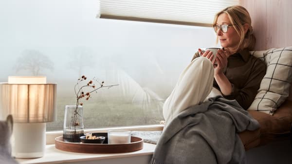 A woman is sitting in a window seat enjoying a break with a cup of tea. On the window ledge is a SYMFONISK speaker lamp.