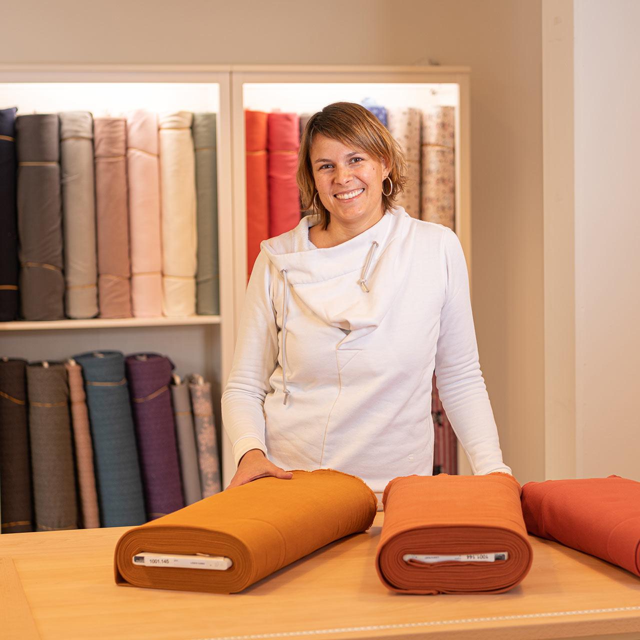 A woman is standing in front of a shelf with various bales of fabric at a counter on which fabrics are lying.