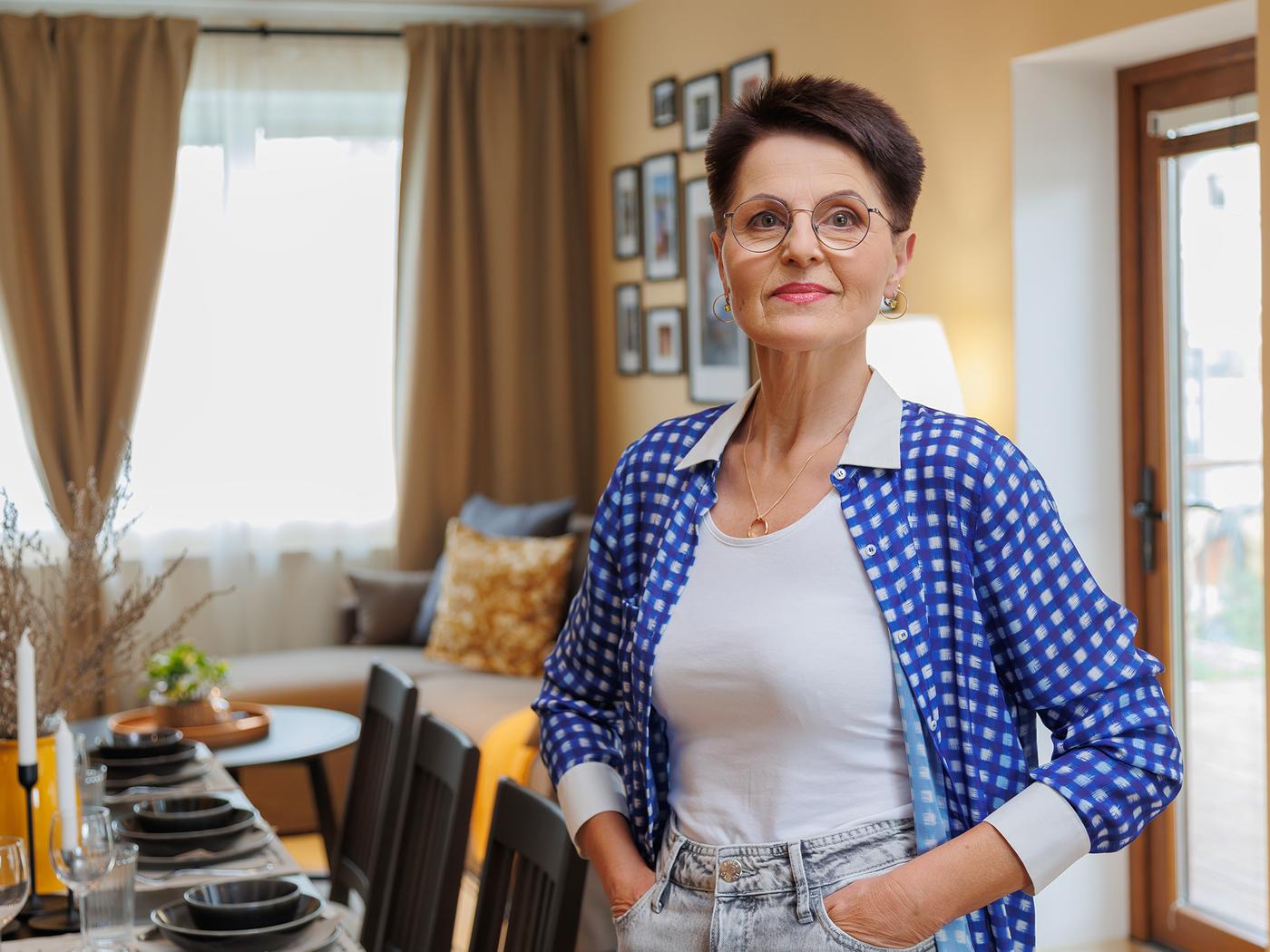 A woman is standing in the living room by the dining table, looking into the camera. In the background, there is a window with curtains and a wall of pictures in frames.