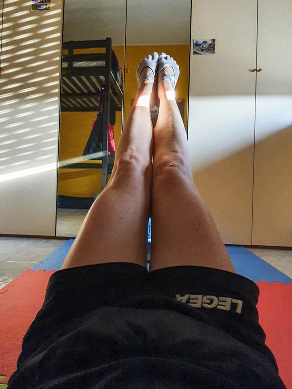 A woman is stretching out her legs, holding them above the floor. She is wearing black exercise shorts and grey yoga socks.