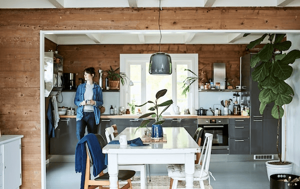 A woman leans on a shiny grey cabinet in a galley kitchen that opens to a dining room with white wood table and chairs.