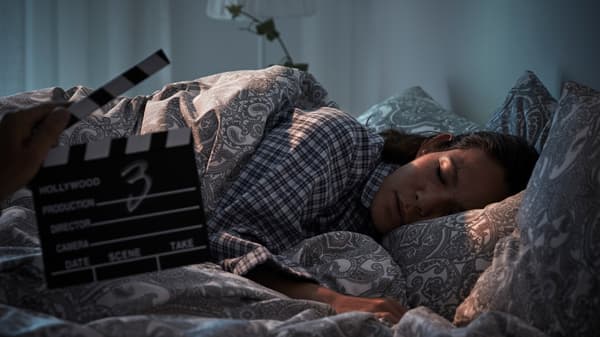 A woman lies in bed as if she is asleep, while another person’s arm holds a director’s clapperboard in front of her.