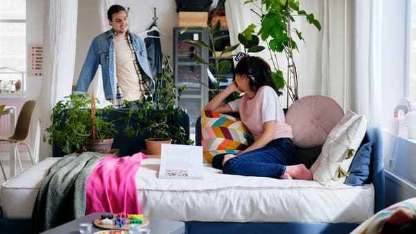 A woman on a blue BLÅKULLEN bed with various colourful cushions and throws, with a man standing behind by some plants.