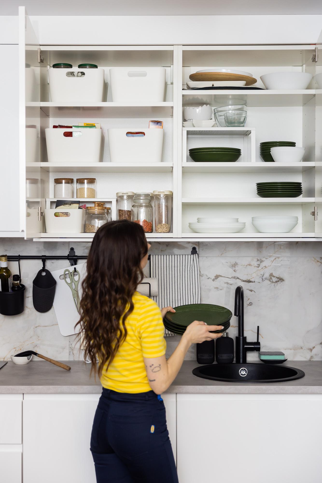 A woman organising kitchen storage using open shelving and containers, demonstrating practical and space-efficient storage solutions.