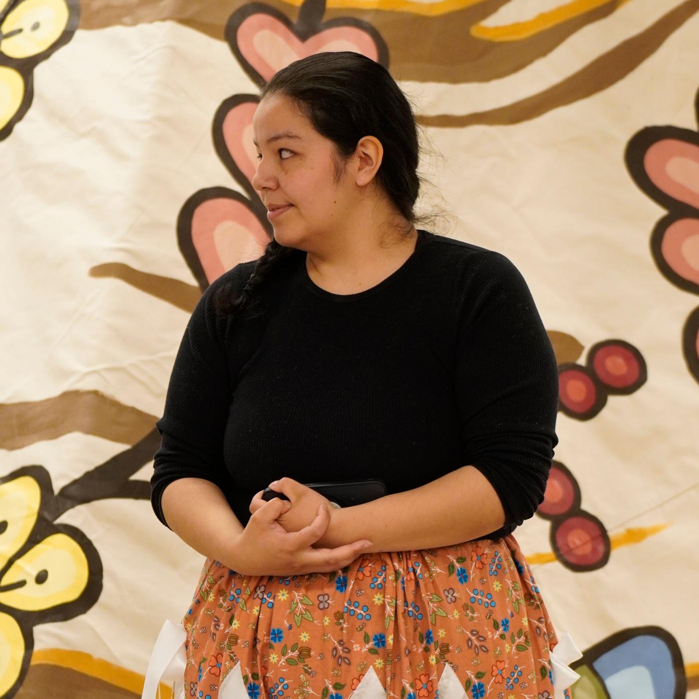 A woman outfitted in a long skirt is looking to the side while standing in front of a tipi.