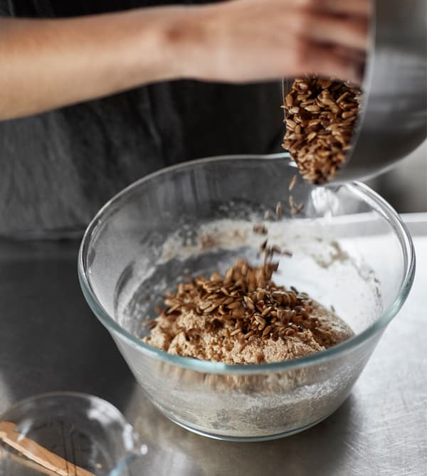 A woman pours roasted sunflower seeds into a glass bowl with dough.