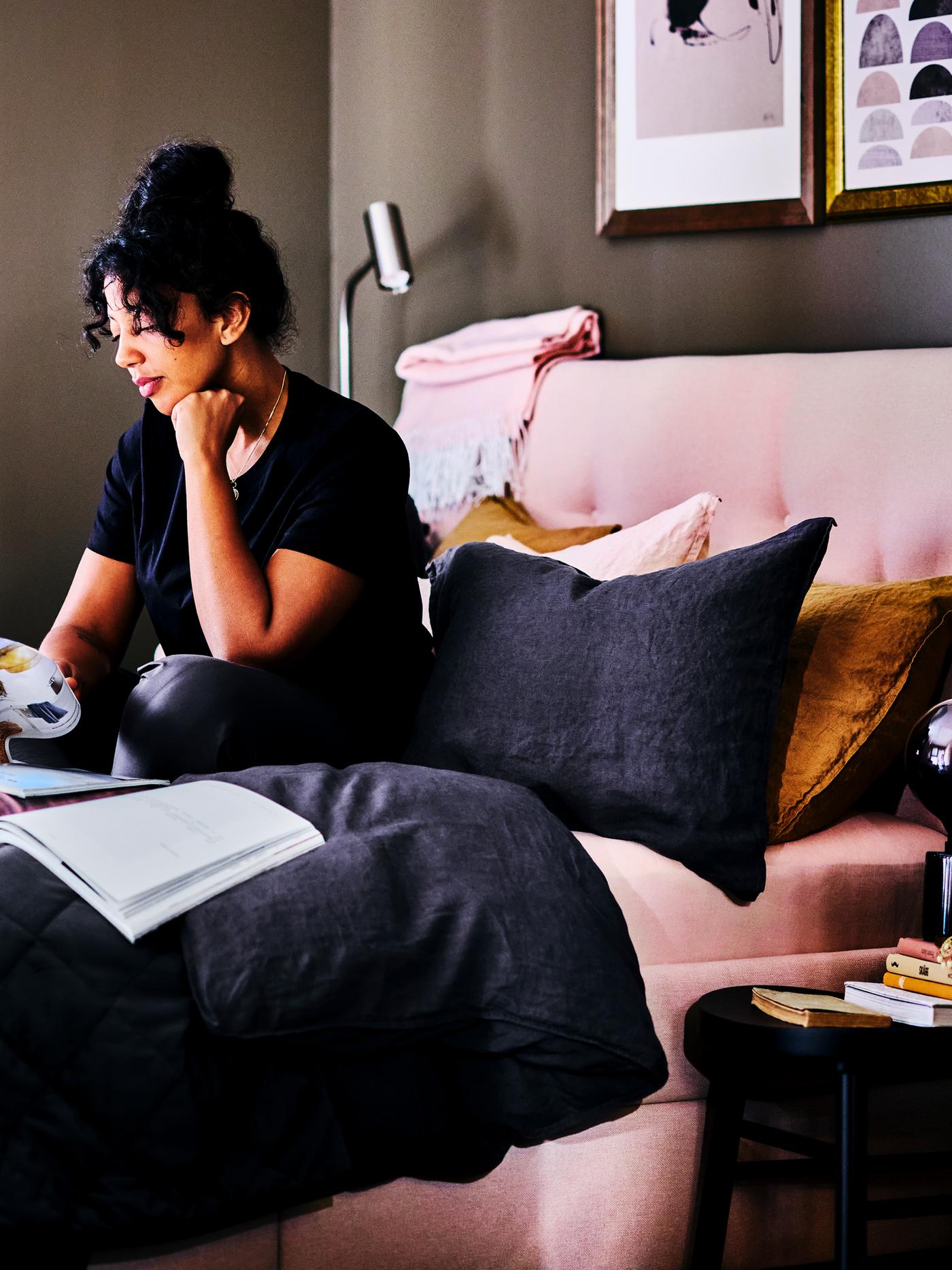 A woman reading a magazine while sitting under the covers in a light pink IDANÄS upholstered storage bed.