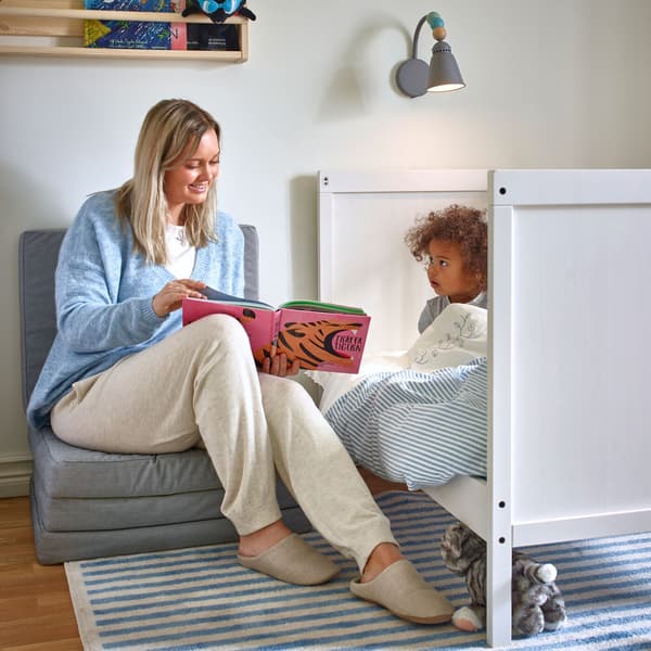 A woman, seated on a foldable IKEA SLÄKT light grey mattress, with an open book, reading to a small child in bed.