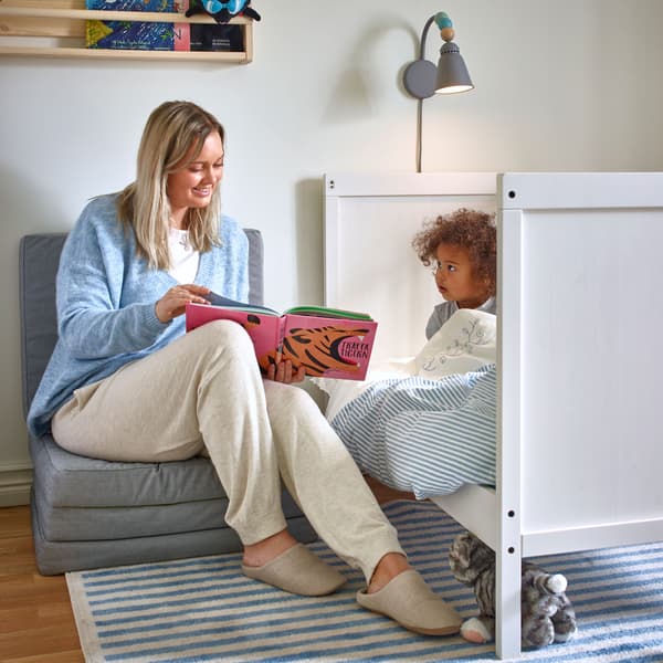 A woman, seated on a foldable IKEA SLÄKT light grey mattress, with an open book, reading to a small child in bed.