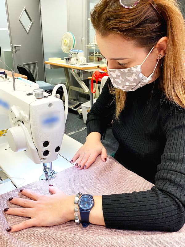 A woman sits at a sewing machine wearing a mouth guard and sews an old rose-coloured curtains