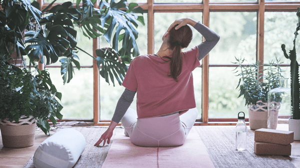A woman sits cross-legged on a VÅRDANDE yoga rug facing a window, stretching her neck in a peaceful space with plants.