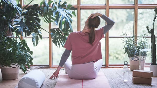 A woman sits cross-legged on a VÅRDANDE yoga rug facing a window, stretching her neck in a peaceful space with plants.