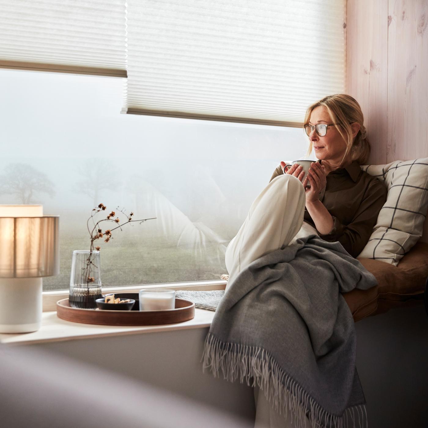 A woman sits on a broad window sill and looks out at a misty day, while rugged up with a blanket, cushion and cup of tea.