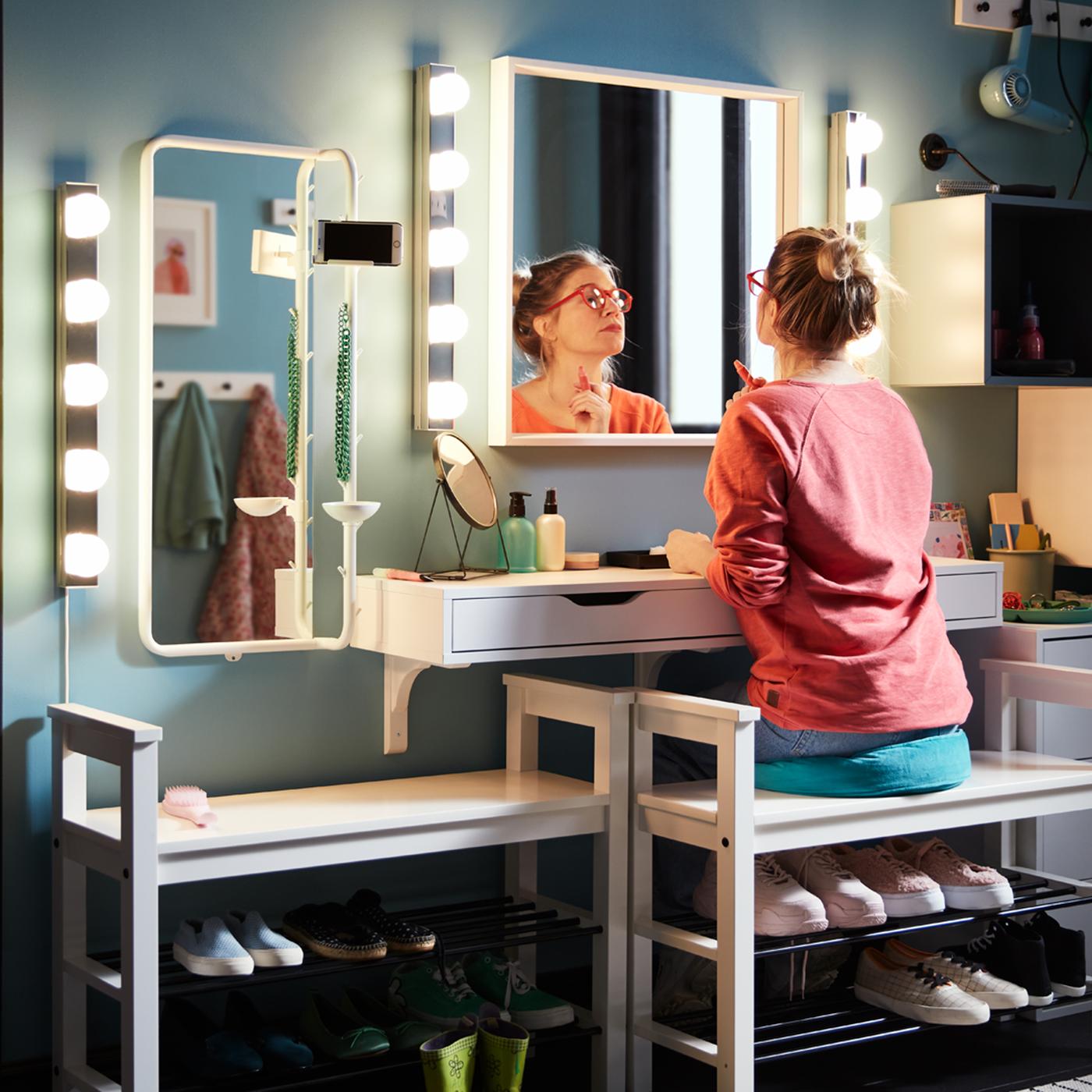 A woman sits on a cushion on a HEMNES bench with shoe storage and does her make-up in front of a NISSEDAL mirror.