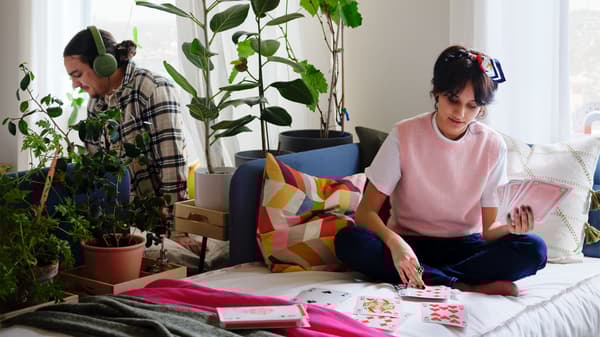 A woman sits on a medium-blue BLÅKULLEN bed. Behind a room-dividing combo of a plant swath and a curtain sits a man.
