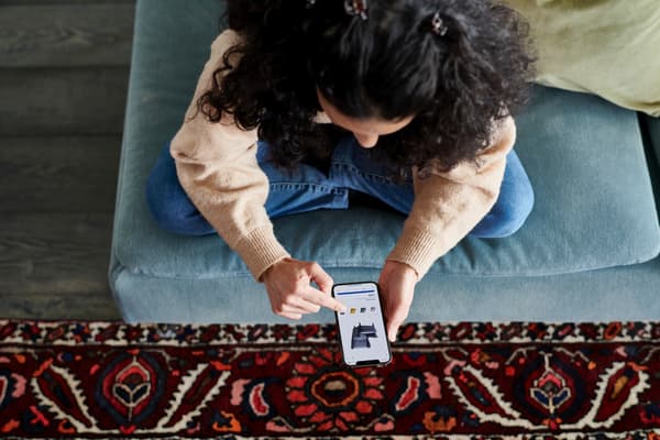 A woman sits on a stool with her legs crossed, checking product availability for an armchair in the IKEA app. 