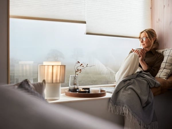 A woman sitting in a cosy window seat with soft lighting, wrapped in a blanket beside a lamp and wooden tray.