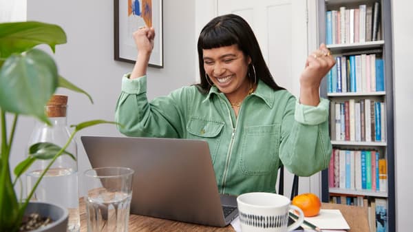 A woman sitting in front of a laptop is celebrating with her hands in the air.