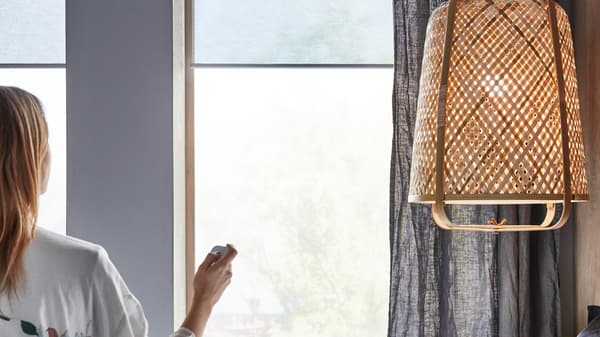 A woman, sitting near a KNIXHULT pendant lamp, uses a remote control on KADRILJ roller blinds hanging in the windows.
