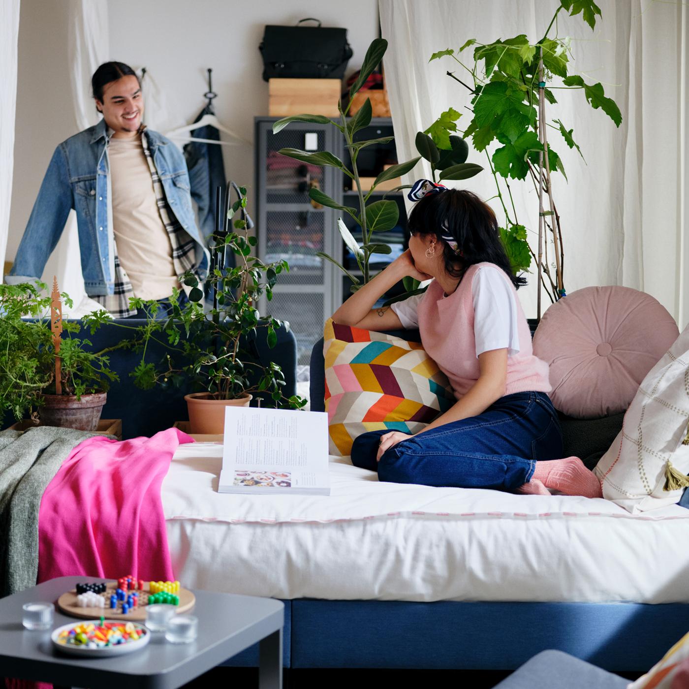 A woman sitting on a BLÅKULLEN upholstered bed frame with scatter cushions looking at a man standing next to an IVAR cabinet.