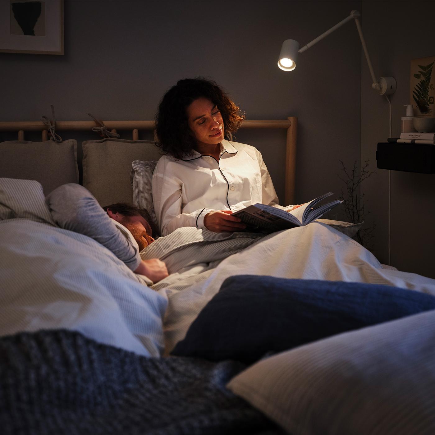 A woman sitting up in bed, reading a book under a reading lamp, with a man lying down in bed beside her.