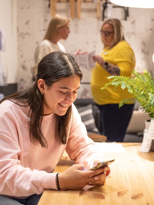 A woman smiles and looks at her mobile, while two customers in the background are looking at a buying guide.