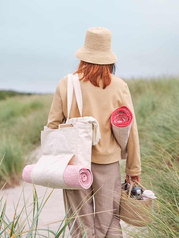 A woman stands among grassy dunes with a VÅRDANDE canvas bag slung over her shoulder, carrying a jute basket and throw.