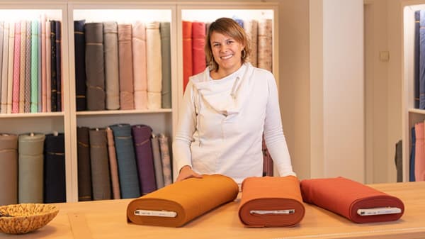 A woman stands in front of a shelf with bales of fabric at a worktable covered with various fabrics.